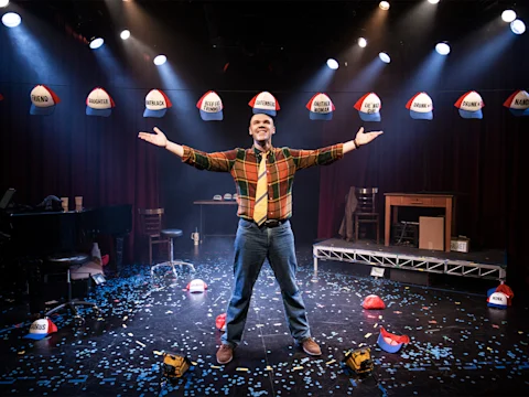 A man stands center stage with arms outstretched, surrounded by hard hats labeled with U.S. state names, under spotlights and scattered confetti.