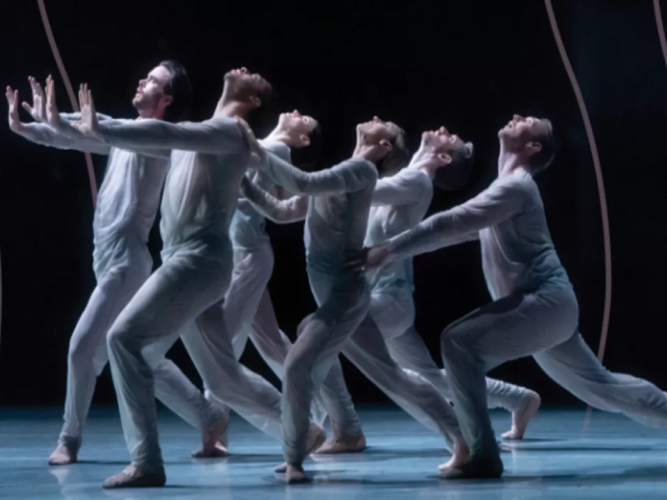 Dancers in white costumes perform a synchronized routine on stage, with dramatic arm placements and poised stances, set against a dark background.