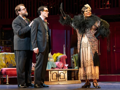 Three actors in vintage costumes perform on stage; two men in suits stand together while a woman in a feathered robe raises her gloved hand in a dramatic gesture.