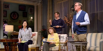 Four adults are engaged in conversation in a well-furnished living room, with one sitting on a couch, one on a chair, and two standing. Walls are decorated with framed pictures.