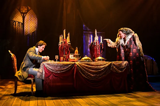 A man in a suit eats at a long table while a cloaked figure points at him with a lit candle.