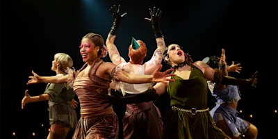 Five performers in colorful costumes and dramatic makeup strike a theatrical pose on stage with arms outstretched, under focused stage lighting.