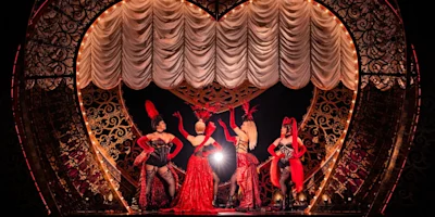 Four performers in elaborate red costumes and headpieces pose on stage in front of a large heart-shaped set with gold accents and draped curtains.
