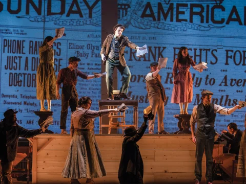 A group of actors on stage performing a musical, dressed in period costumes. They hold newspapers and stand on various elevated platforms. A large, projected newspaper backdrop is in the background.