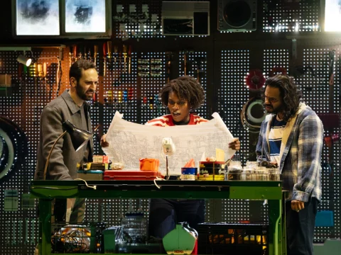 Three people stand around a workbench examining a large blueprint. The background has various tools and equipment displayed on a pegboard.