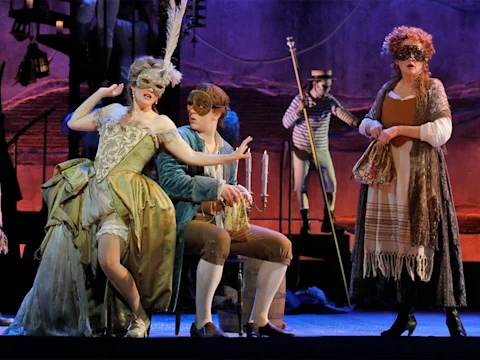 Three people in elaborate period costumes and masks perform on stage; one sits holding a candlestick, while two stand, with a fourth person in striped clothing in the background.