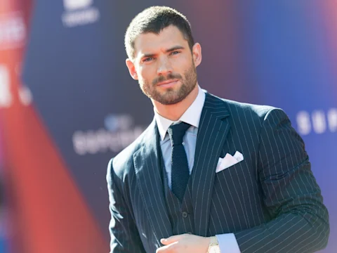 A man with short dark hair and a beard wearing a pinstripe suit, dress shirt, and tie, stands outdoors against a blurred background.