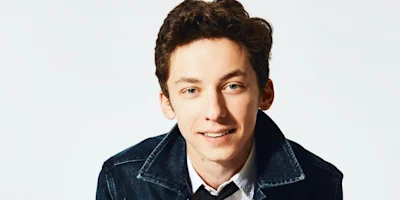 A young man with short curly brown hair, wearing a white shirt and dark denim jacket, smiles at the camera against a plain light background.