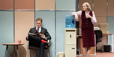 A man in a suit reads a magazine while seated; a woman in office attire leans on a water cooler, looking at him. Both are in a retro-style office setting.