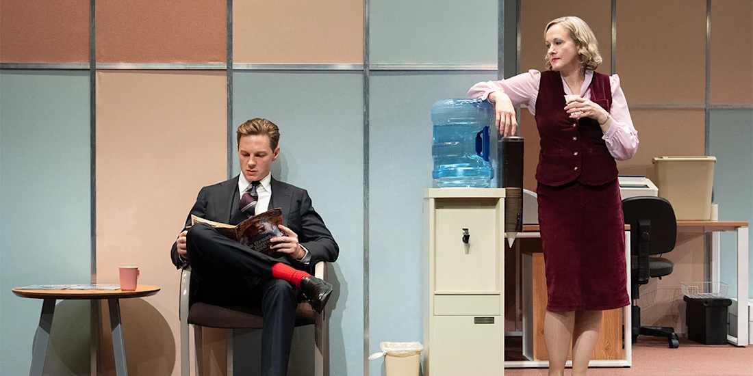 A man in a suit reads a magazine while seated; a woman in office attire leans on a water cooler, looking at him. Both are in a retro-style office setting.