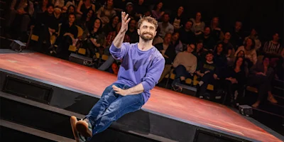 A man in a purple sweater and jeans sits on the edge of a stage, gesturing with his hand, while an audience watches in the background.