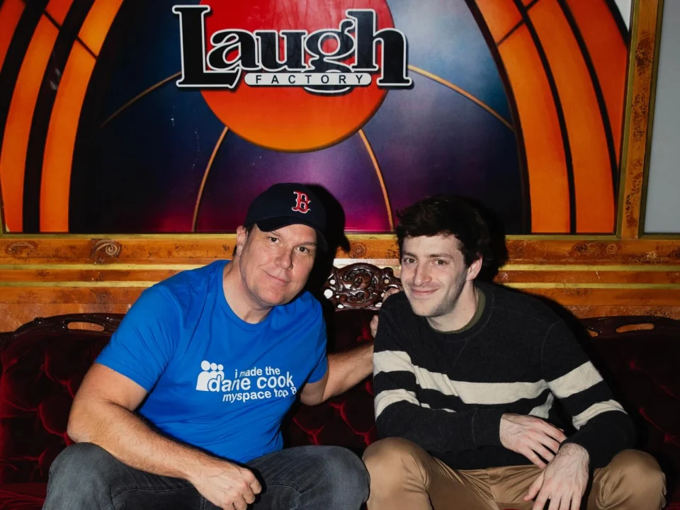 Two people sitting on a red couch in front of a 'Laugh Factory' sign. The person on the left wears a blue t-shirt and a black cap, and the person on the right wears a striped sweater.