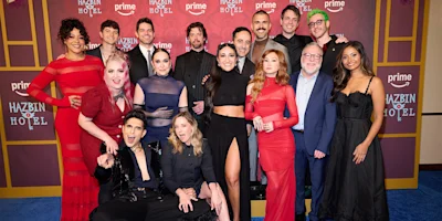 A group of people pose together on a blue carpet in front of a Hazbin Hotel step-and-repeat backdrop at a Prime Video event.