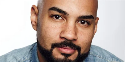 A close-up portrait of a man with a shaved head and a beard wearing a denim shirt.