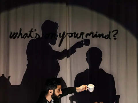 A person holding a cup near a projected image of two silhouettes, one asking "what's on your mind?" while holding a cup above the other seated silhouette.