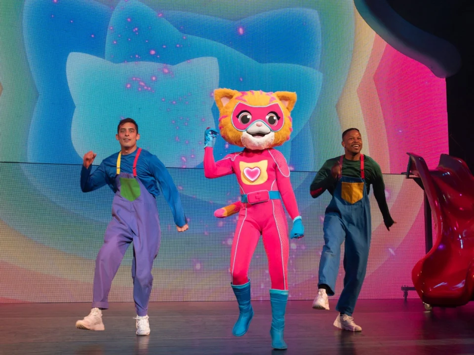 Three performers are on stage; one is dressed in a pink superhero cat costume, while the other two wear colorful outfits. The background features bright, colorful designs.