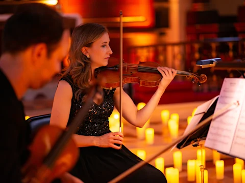 A woman in a black dress plays the violin in a candlelit setting, accompanied by another violinist in the foreground.
