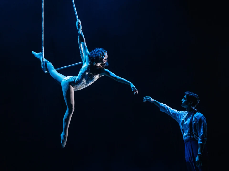 Aerial performer in a bodysuit is suspended on a rope, reaching toward a standing man in a dimly lit setting.