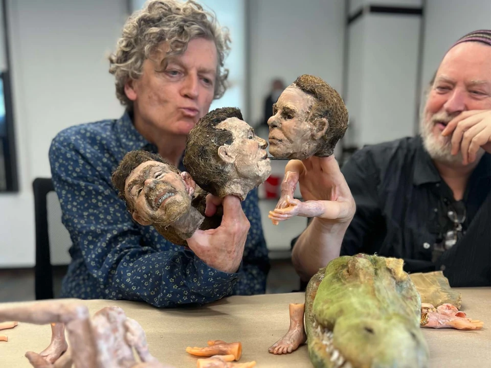 Two people are seated at a table handling art pieces, including three sculpted heads with lifelike faces and a green crocodile sculpture. Various small sculpted body parts are also on the table.