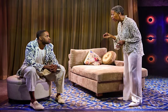 Two people are having a serious conversation in a living room setting with patterned furniture, draped curtains, and a blue-patterned rug.