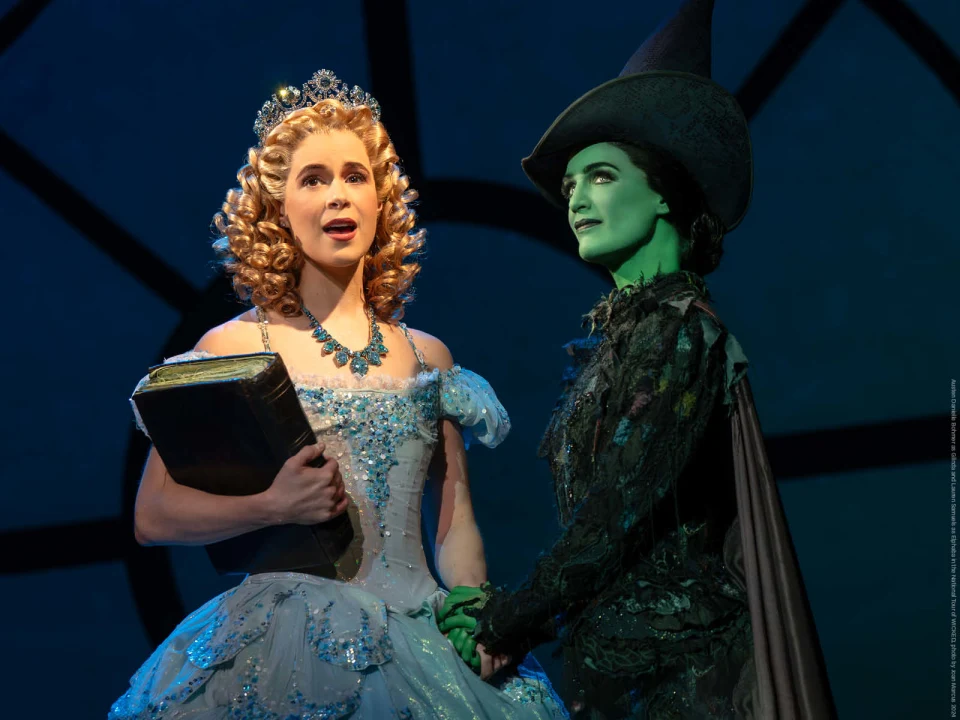 Production photo of WICKED in San Francisco, showing actors on stage, one dressed in a sparkling blue gown and crown, the other in a green face and dark attire.