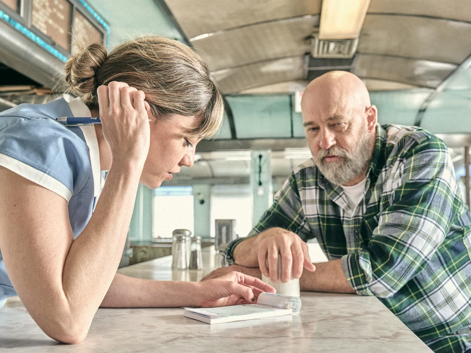 A waitress in a blue uniform is taking an order from a bearded, bald man in a green plaid shirt at a diner counter.