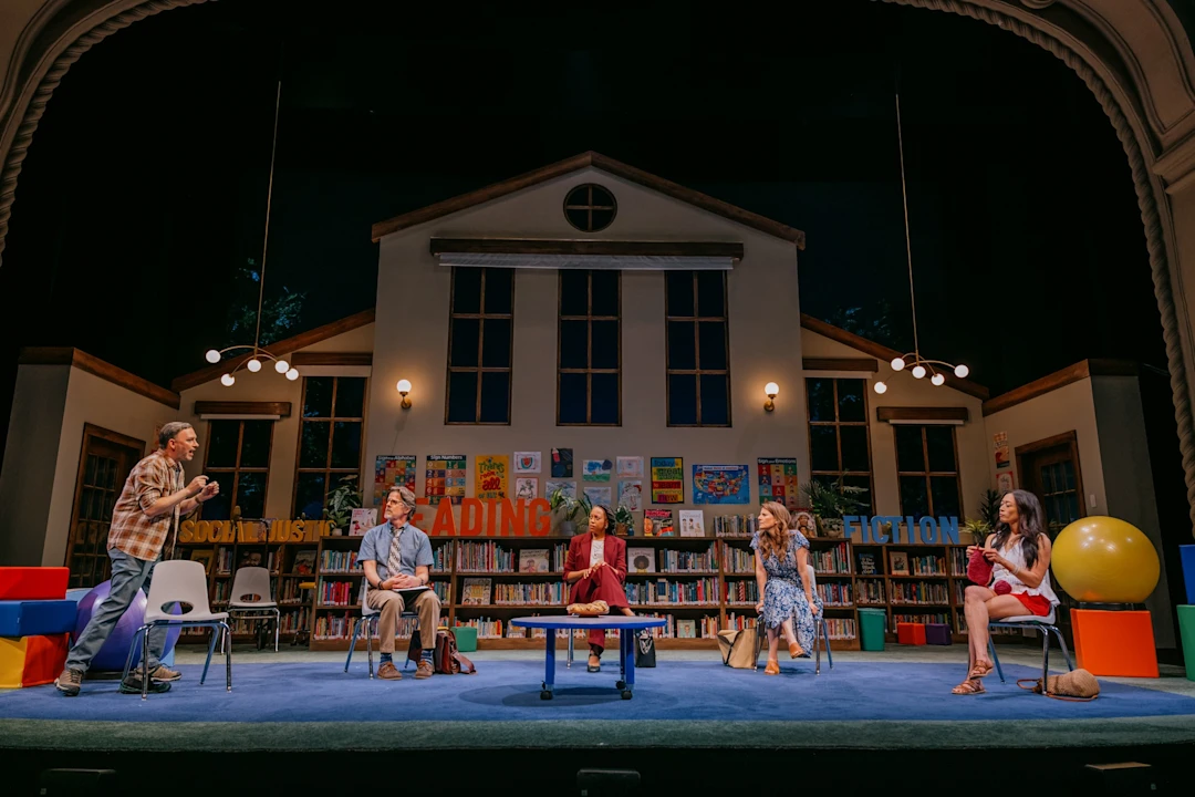 Five people sit and converse on a stage set resembling a library, with bookshelves, large letters spelling "READING" and "FICTION," and colorful props visible in the background.