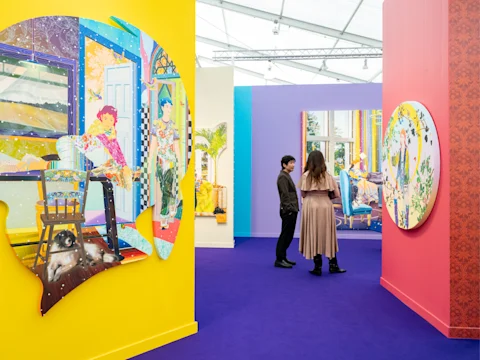 Two people view colorful, contemporary art pieces displayed on vibrant yellow and red walls in a brightly lit gallery space with a purple floor.