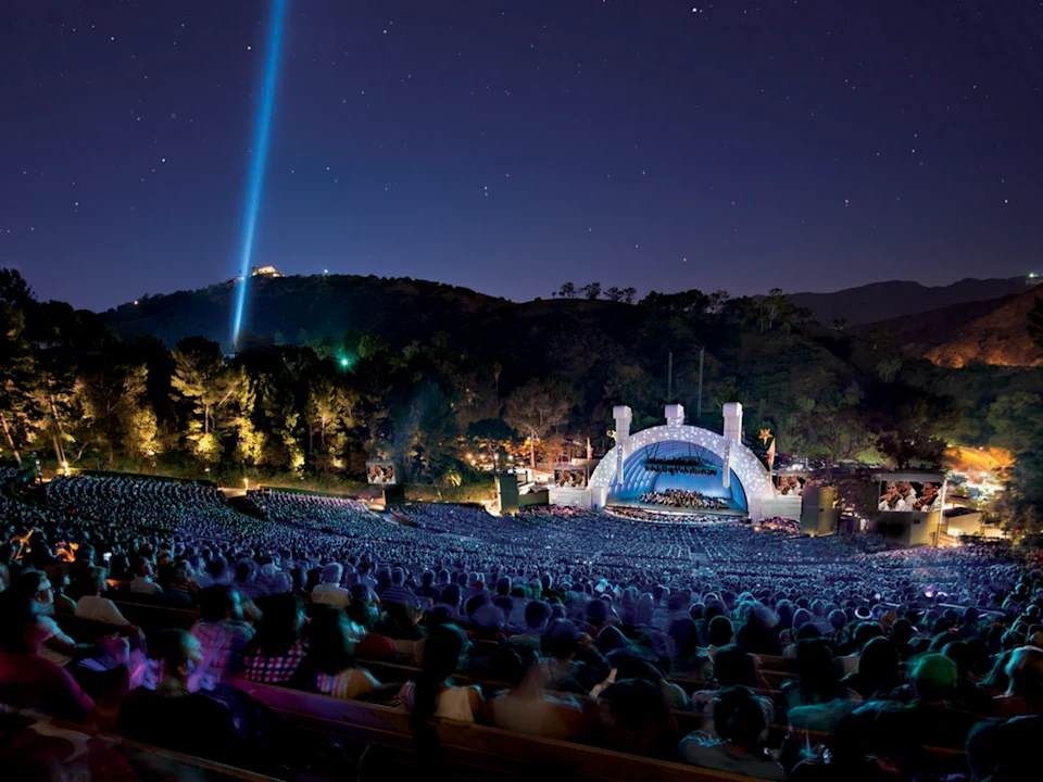 Outdoor amphitheater at night with a crowd watching a performance; stage lit up, hills and a beam of light in the background.