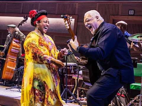 A woman in a yellow floral dress sings while a man plays guitar next to her on stage. Other musicians play instruments in the background.