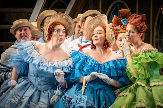 A group of people in colorful period costumes and bonnets, some women in blue and one in green, appear to converse during a theatrical performance—a production shot of HMS Pinafore with the ENO Chorus captured by Marc Brenner.