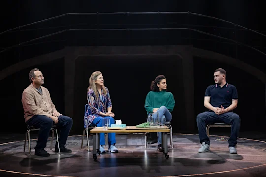 Four people sit on chairs in a semicircle on a stage, with a coffee table in front of them holding tissues, glasses, and a jug of water.