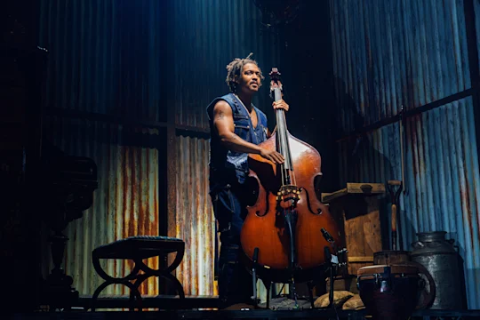A musician stands on stage playing a double bass, surrounded by rustic props and dramatic lighting.