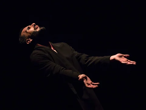 A man in a dark suit stands against a black background with his head tilted back and arms outstretched, palms facing up.