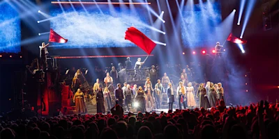 A large cast performs on stage with dramatic lighting; two people wave red flags, while others sing in front of a backdrop with blue and white light effects.