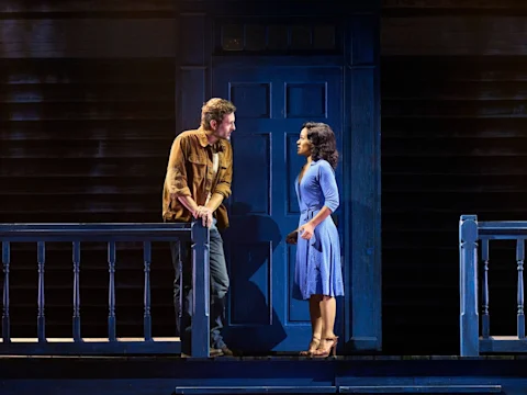 Production photo of The Notebook in Costa Mesa, showing two actors perform on stage; a man leans on a porch railing while a woman in a blue dress stands facing him in front of a blue door and dark house backdrop.