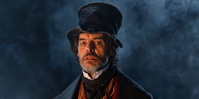 A man with a beard and mustache wearing a tall, crumpled hat and period clothing stands against a smoky, dark background.