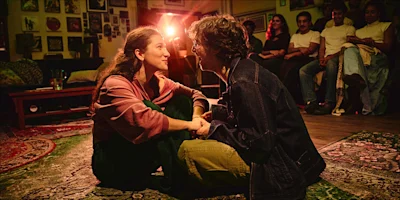 Two people sit facing each other and hold hands on a living room floor while a small audience watches from the background.