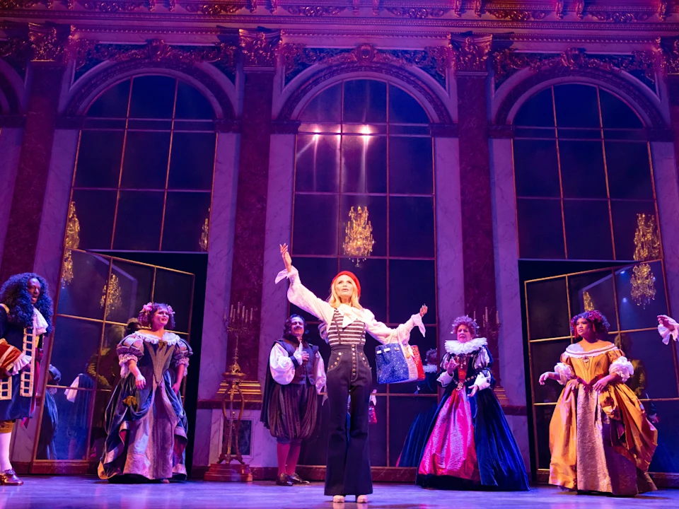 Production shot of The Queen of Versailles in New York, with Kristin Chenoweth as Jackie Siegel and a group of actors in elaborate period costumes performs on a stage with tall windows and chandeliers; one stands at the center gesturing dramatically.