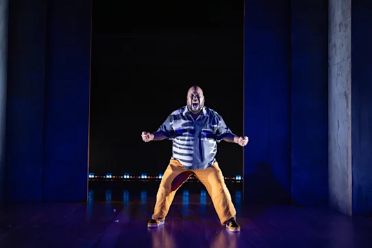 A man stands center stage under dramatic lighting, wearing an untucked shirt and orange pants, with his arms bent and mouth open as if shouting or singing.