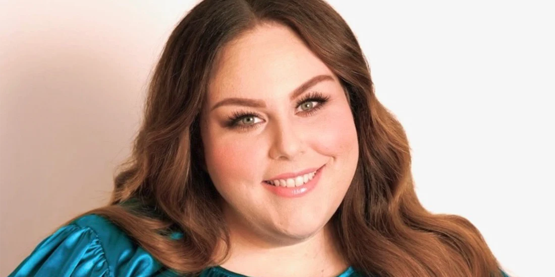 A woman with long brown hair and light makeup smiles at the camera, wearing a teal top against a plain light background.