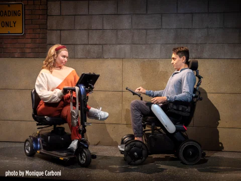 Two individuals in motorized wheelchairs facing each other against a brick wall. The person on the left is wearing a white and beige sweater, and the person on the right is wearing a gray shirt. Photo by Monique Carboni.