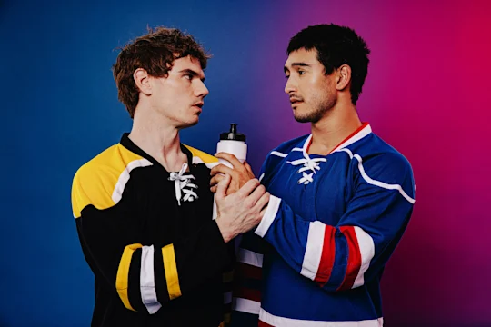 Two male hockey players in different jerseys stand close, both holding a water bottle, looking seriously at each other against a blue and pink gradient background.