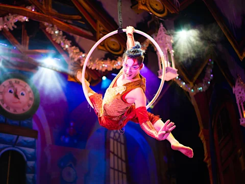 Production photo of Cirque Dreams Holidaze in New Brunswick, showing a circus performer in a red and gold costume poses mid-air on a suspended hoop, with colorful stage lights and a decorative background behind them.