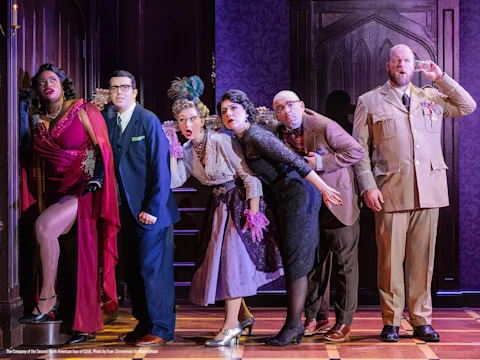 Six actors dressed in vintage costumes lean toward a door with surprised and curious expressions on a theatrical stage set.