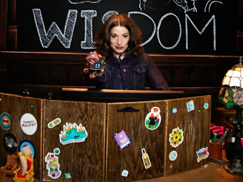 Production photo of Madelyn Murphy, stands behind a wooden desk decorated with stickers, holding a large 20-sided die in front of a blackboard.