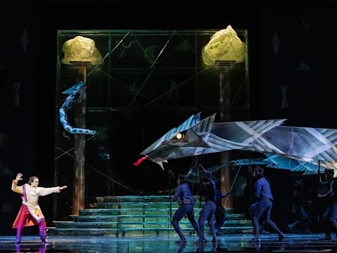 Production photo of The Magic Flute—Holiday Presentation in New York, showing a stage performance features a man in a white costume confronting a large, dragon-like puppet operated by several people against a backdrop of stone pillars and stairs.