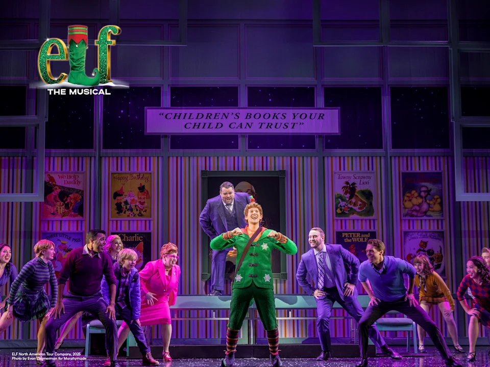 Stage scene from “Elf The Musical” featuring a group of actors in colorful costumes, with one actor dressed as an elf in the center, set against a backdrop of children’s books.