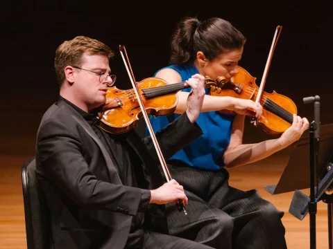 Two musicians play violins on stage, both wearing formal attire. One wears a black suit and glasses, the other wears a blue blouse.