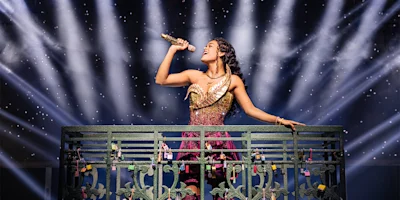 A woman in a sparkling dress sings into a gold microphone on a balcony, with bright stage lights and a starry backdrop behind her.